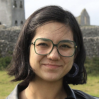 The image features a young woman with short black hair,
wearing green-framed glasses and large circular earrings.
They have a gentle smile and are dressed in a black leather
jacket. The background showcases an old stone structure
with greenery, suggesting a historic or rural setting. The lighting is natural, giving the photo a
warm and candid feel.